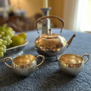 May include: A silver teapot, creamer, and sugar bowl set on a blue tablecloth. The teapot has a classic design with a rounded body and a spout. The creamer and sugar bowl are both small and round.