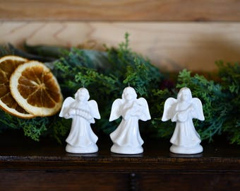 May include: Three white ceramic angel figurines with outstretched wings, standing on a dark wooden surface. The angels are in front of a backdrop of greenery and dried orange slices, creating a festive, decorative display.