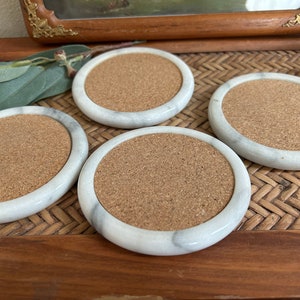 Pu&ograve; includere: Quattro sottobicchieri in marmo bianco con inserti in sughero. I sottobicchieri sono rotondi e hanno un bordo leggermente rialzato. Sono disposti su una superficie in vimini intrecciato.