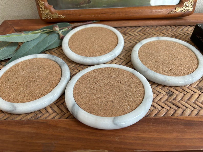 Pu&ograve; includere: Quattro sottobicchieri in marmo bianco con inserti in sughero. I sottobicchieri sono rotondi e hanno un bordo leggermente rialzato. Sono disposti su una superficie in vimini intrecciato.