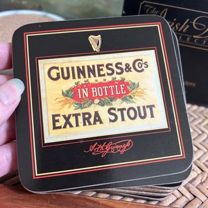 Pu&ograve; includere: Un set di sottobicchieri neri con bordo dorato che presenta un'etichetta vintage di Guinness con il testo "GUINNESS & Co's IN BOTTLE EXTRA STOUT" e una firma che recita "Arthur Guinness".