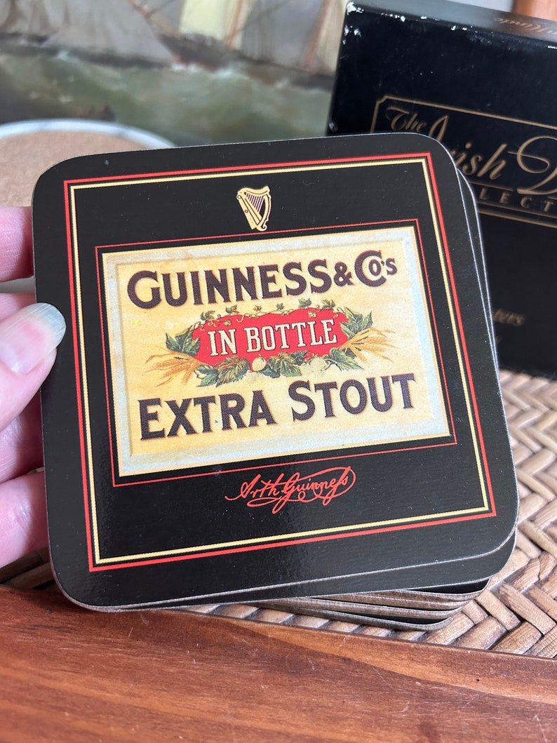 Pu&ograve; includere: Un set di sottobicchieri neri con bordo dorato che presenta un'etichetta vintage di Guinness con il testo "GUINNESS & Co's IN BOTTLE EXTRA STOUT" e una firma che recita "Arthur Guinness".