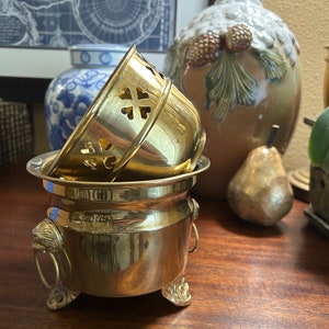 May include: A set of two gold-toned brass plant pots with decorative lion head handles and feet. The smaller pot has a decorative cut-out design.