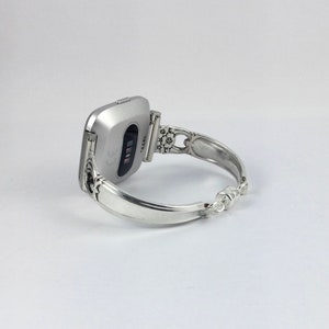 May include: A silver-toned fitness tracker with a black screen is attached to a silver bracelet with an ornate design.