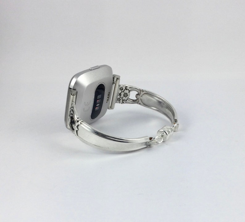 May include: A silver-toned fitness tracker with a black screen is attached to a silver bracelet with an ornate design.