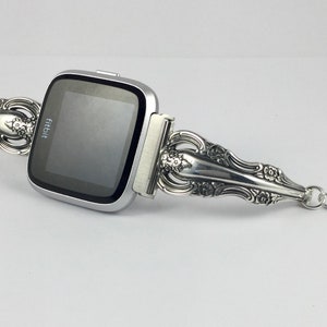 May include: A silver Fitbit smartwatch with a custom-made band crafted from a vintage silverware piece. The band features intricate floral designs and a small charm attached to the end.