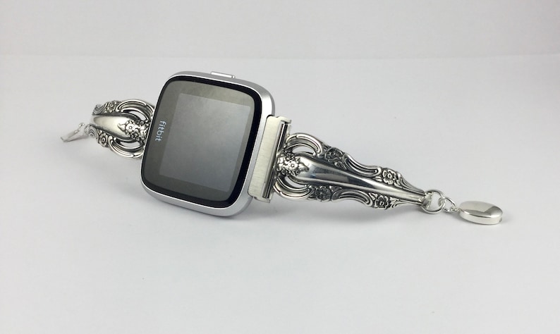 May include: A silver Fitbit smartwatch with a custom-made band crafted from a vintage silverware piece. The band features intricate floral designs and a small charm attached to the end.