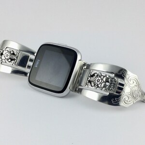 May include: A silver Fitbit smartwatch with a decorative, ornate, silver band made from a repurposed utensil. The band has a floral design.