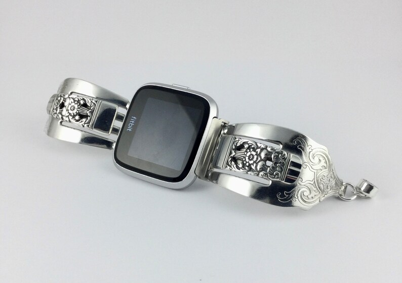 May include: A silver Fitbit smartwatch with a decorative, ornate, silver band made from a repurposed utensil. The band has a floral design.