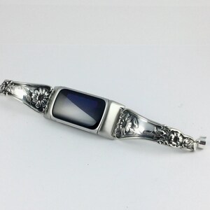 May include: A silver-toned watch band with floral and scrollwork detailing. The band features a rectangular, dark-colored screen in the center. The design includes ornate embellishments and a clasp.