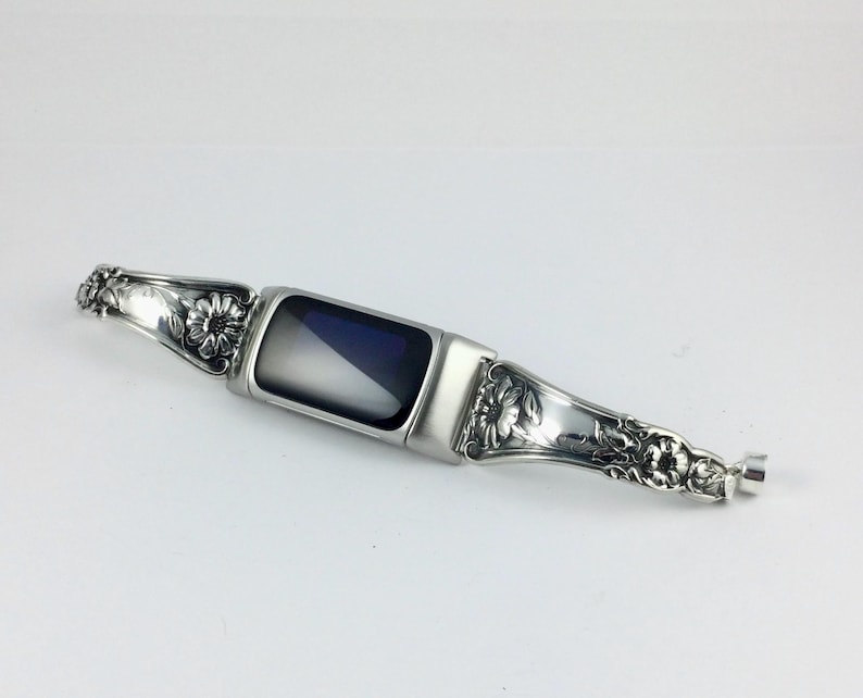 May include: A silver-toned watch band with floral and scrollwork detailing. The band features a rectangular, dark-colored screen in the center. The design includes ornate embellishments and a clasp.
