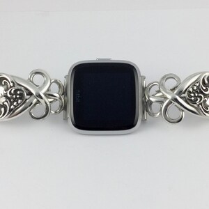 May include: Silver watch band made from repurposed silverware with a black Fitbit watch face. The band features a decorative floral design.
