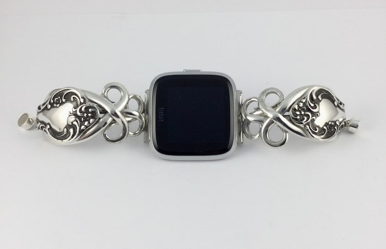 May include: Silver watch band made from repurposed silverware with a black Fitbit watch face. The band features a decorative floral design.