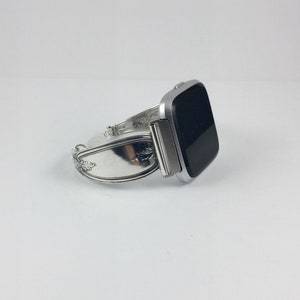 May include: A silver watch band made from a repurposed silverware piece. The band is designed to fit a square-shaped smartwatch. The band has a decorative floral design.