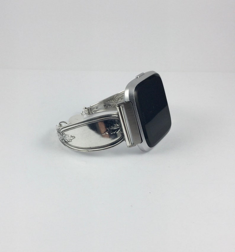 May include: A silver watch band made from a repurposed silverware piece. The band is designed to fit a square-shaped smartwatch. The band has a decorative floral design.