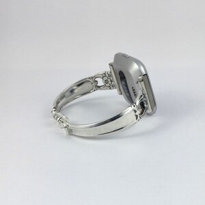May include: A silver-toned fitness tracker attached to a silver-toned silverware bracelet. The bracelet is made from a bent piece of silverware with a decorative design.