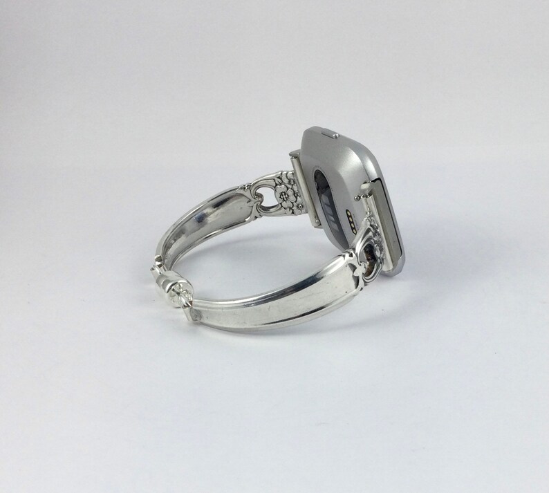 May include: A silver-toned fitness tracker attached to a silver-toned silverware bracelet. The bracelet is made from a bent piece of silverware with a decorative design.