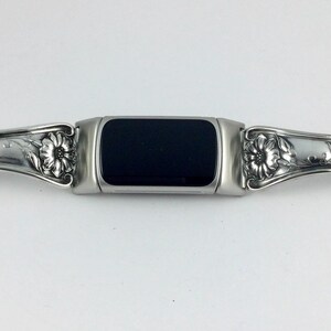 May include: A silver-toned watch band with a black rectangular center. The band features ornate floral and leaf designs on each side. The watch band is made of metal and has a vintage or antique aesthetic.