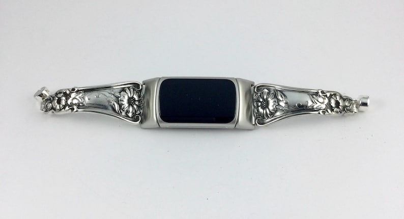 May include: A silver-toned watch band with a black rectangular center. The band features ornate floral and leaf designs on each side. The watch band is made of metal and has a vintage or antique aesthetic.