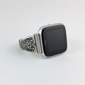 May include: Silver Fitbit Versa smartwatch with a decorative band. The band features an ornate floral design. The watch has a black screen and a silver bezel. The Fitbit logo is visible on the watch face.