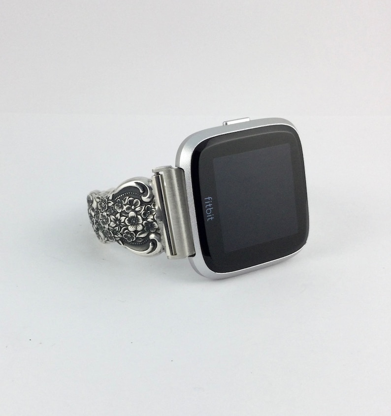 May include: Silver Fitbit Versa smartwatch with a decorative band. The band features an ornate floral design. The watch has a black screen and a silver bezel. The Fitbit logo is visible on the watch face.