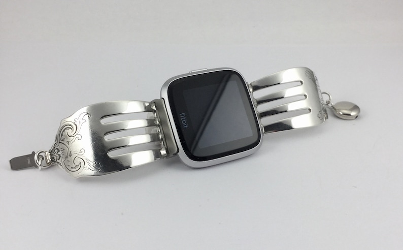 Pu&ograve; includere: Un orologio intelligente Fitbit argentato con uno schermo nero &egrave; attaccato a un braccialetto argentato realizzato con forchette riutilizzate. Il braccialetto ha un ciondolo decorativo.