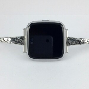 May include: A silver Fitbit smartwatch with a black screen and a decorative silver band with floral designs.