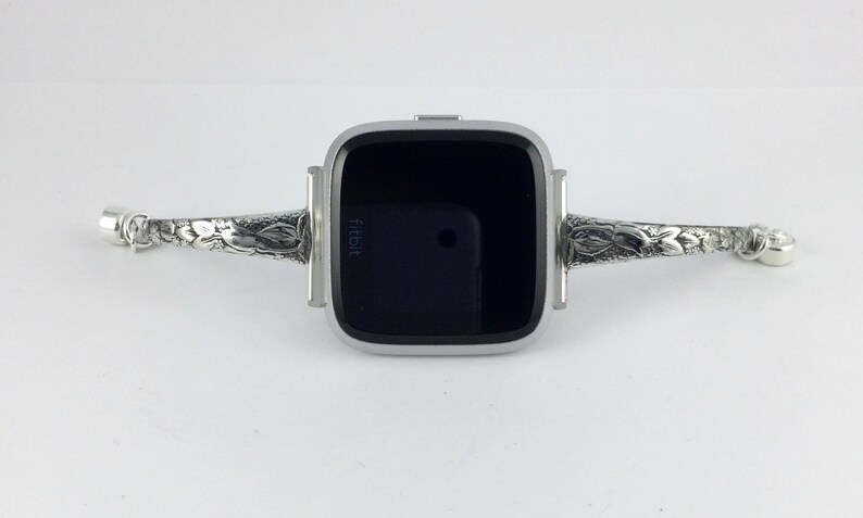 May include: A silver Fitbit smartwatch with a black screen and a decorative silver band with floral designs.