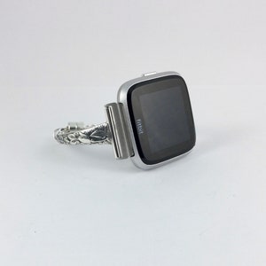 May include: A silver Fitbit smartwatch with a black screen and the word "Fitbit" displayed on the screen. The watch is attached to a silver metal band with a decorative design.