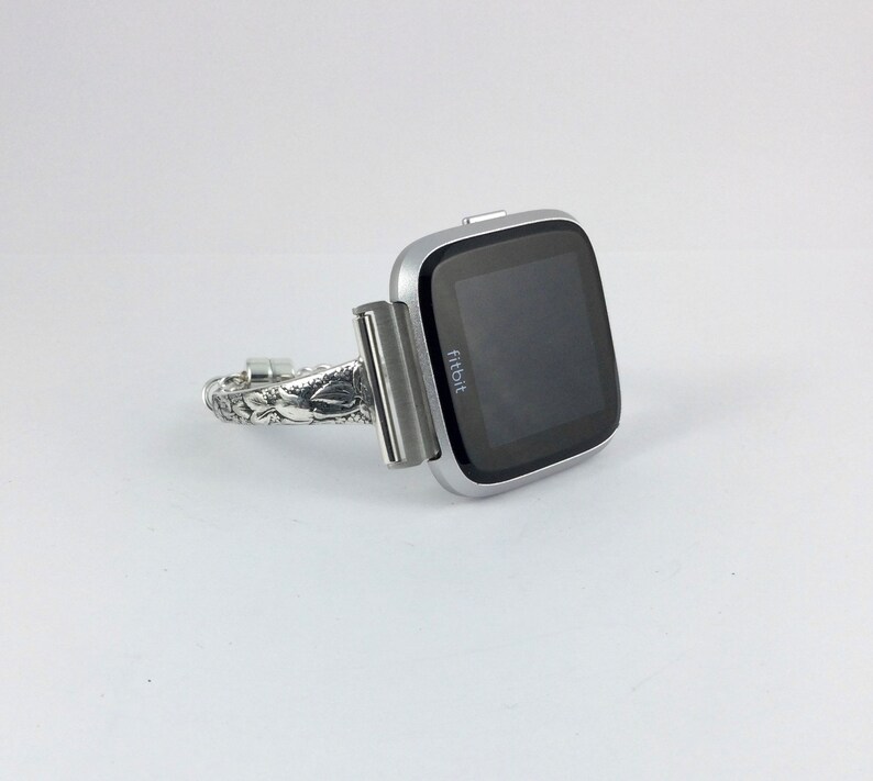 May include: A silver Fitbit smartwatch with a black screen and the word "Fitbit" displayed on the screen. The watch is attached to a silver metal band with a decorative design.