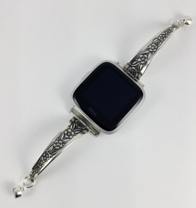 Puede incluir: Pulsera de reloj plateada con un dise&ntilde;o floral. La pulsera est&aacute; unida a un reloj inteligente cuadrado con una pantalla negra y la palabra "fitbit" impresa en ella.