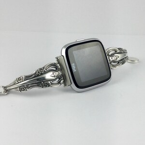 May include: A silver Fitbit smartwatch with a silver, ornate, spoon-shaped band. The band is decorated with floral designs. The watch face displays the word "fitbit".