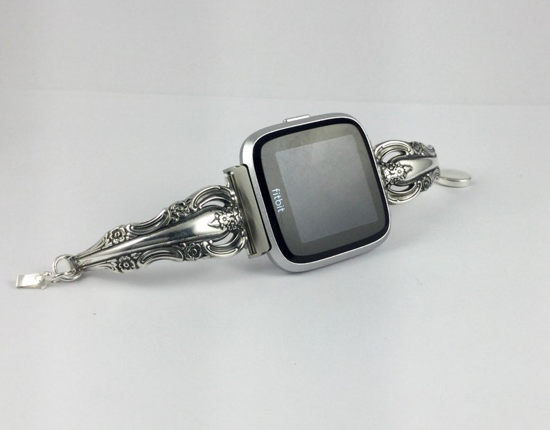 May include: A silver Fitbit smartwatch with a silver, ornate, spoon-shaped band. The band is decorated with floral designs. The watch face displays the word "fitbit".