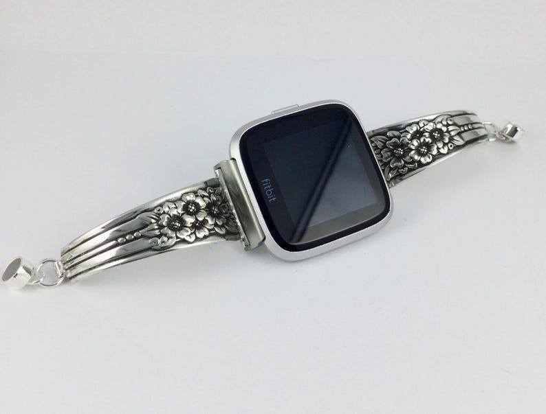 Pu&ograve; includere: Cinturino per orologio in argento con un design floreale. Il cinturino &egrave; attaccato a uno smartwatch Fitbit argentato con uno schermo nero.