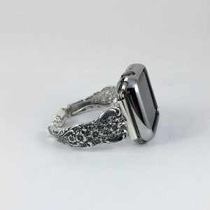May include: Silver-toned Apple Watch band with an ornate floral design. The band features a detailed pattern of flowers and leaves, and the watch face is a sleek, black rectangle. The band is made of metal and has a vintage aesthetic.