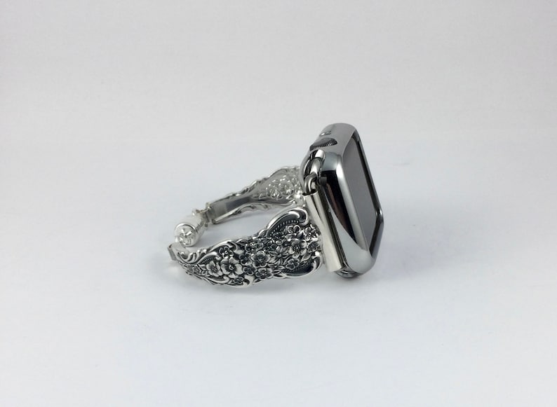 May include: Silver-toned Apple Watch band with an ornate floral design. The band features a detailed pattern of flowers and leaves, and the watch face is a sleek, black rectangle. The band is made of metal and has a vintage aesthetic.
