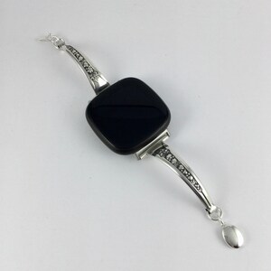 Puede incluir: Un brazalete de plata con una piedra negra cuadrada y un dise&ntilde;o decorativo en la banda.