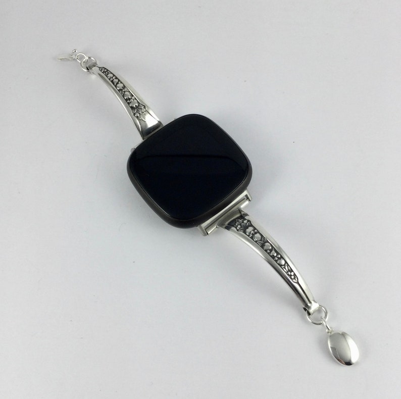 Puede incluir: Un brazalete de plata con una piedra negra cuadrada y un dise&ntilde;o decorativo en la banda.