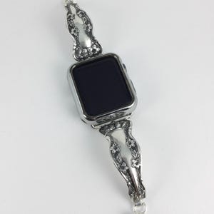 May include: A silver-toned smartwatch featuring a rectangular black screen and a metallic watch band crafted from repurposed silverware handles. The band displays an intricate embossed floral pattern with small blossoms and leaves along the length.