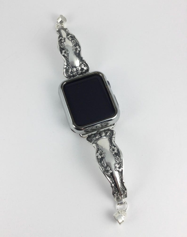 May include: A silver-toned smartwatch featuring a rectangular black screen and a metallic watch band crafted from repurposed silverware handles. The band displays an intricate embossed floral pattern with small blossoms and leaves along the length.