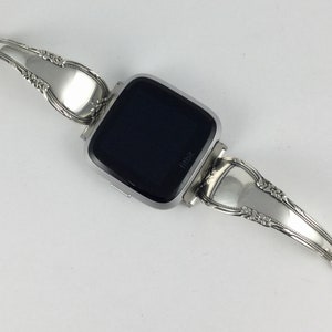 May include: Silver watch band made from repurposed silverware. The band is attached to a silver smartwatch with a black screen that says "Fitbit".