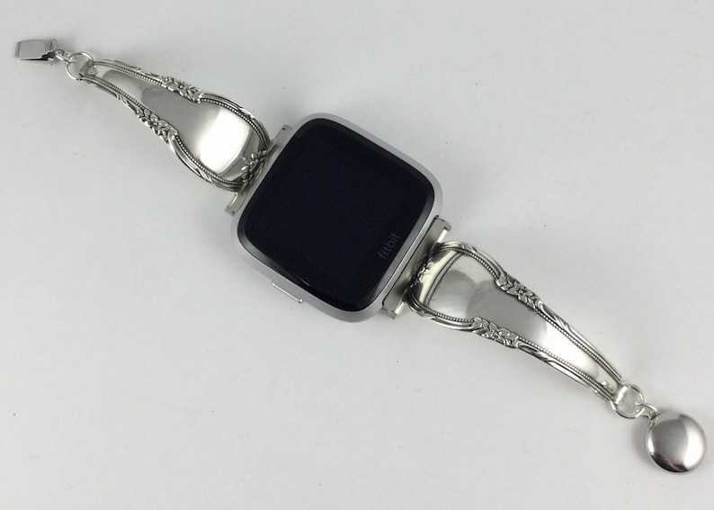 May include: Silver watch band made from repurposed silverware. The band is attached to a silver smartwatch with a black screen that says "Fitbit".