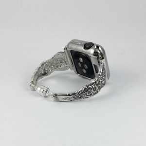 May include: Silver Apple Watch band with an ornate floral design. The band is made of metal and features a magnetic clasp. The watch case is also silver. The band has a vintage, antique aesthetic.