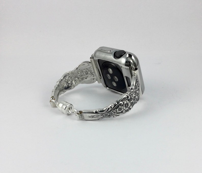 May include: Silver Apple Watch band with an ornate floral design. The band is made of metal and features a magnetic clasp. The watch case is also silver. The band has a vintage, antique aesthetic.