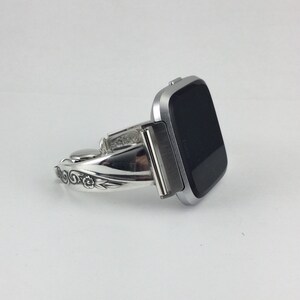 May include: Silver watch band with a floral design. The band is curved and has a silver buckle. The watch face is black and square.