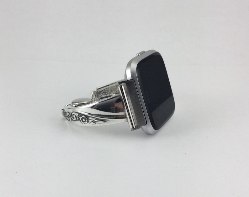 May include: Silver watch band with a floral design. The band is curved and has a silver buckle. The watch face is black and square.