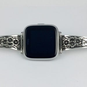 Pu&ograve; includere: Un braccialetto per orologio in argento con un design floreale. Il braccialetto &egrave; composto da due pezzi che si collegano attorno al quadrante dell'orologio. Il quadrante dell'orologio &egrave; nero e ha la scritta "Fitbit" su di esso.