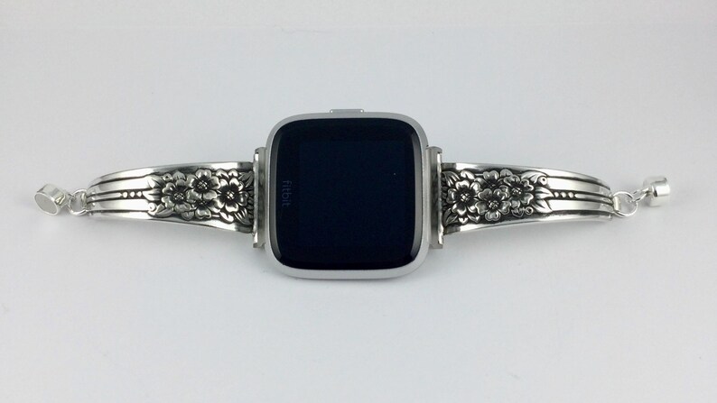 Pu&ograve; includere: Un braccialetto per orologio in argento con un design floreale. Il braccialetto &egrave; composto da due pezzi che si collegano attorno al quadrante dell'orologio. Il quadrante dell'orologio &egrave; nero e ha la scritta "Fitbit" su di esso.