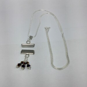 Puede incluir: Collar plateado con un colgante rectangular. El colgante tiene dos barras horizontales y un grupo de peque&ntilde;as cuentas de colores. La cadena es de estilo serpiente delicado, sobre un fondo blanco.
