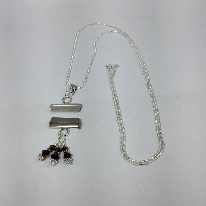 Puede incluir: Collar plateado con un colgante rectangular. El colgante tiene dos barras horizontales y un grupo de peque&ntilde;as cuentas de colores. La cadena es de estilo serpiente delicado, sobre un fondo blanco.